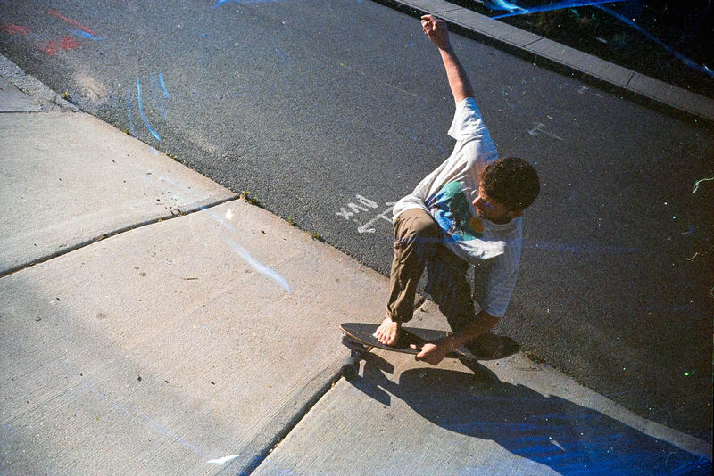 Skateboarder crouched mid-trick on a sidewalk, shot on light-leaked film