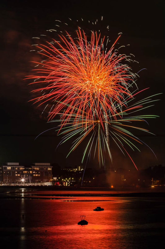 Fireworks bursting over Salem Harbor at night, reflected in the water with boats anchored below