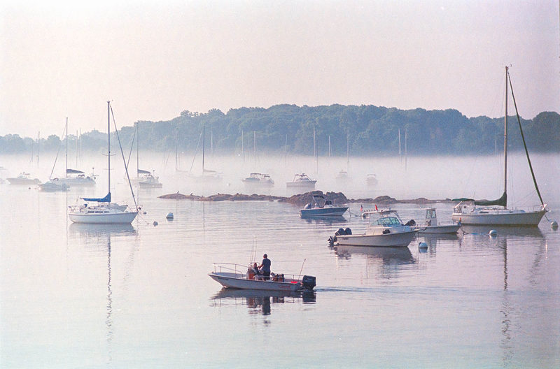 Small fishing boat motoring past moored sailboats in misty morning fog on Salem Harbor