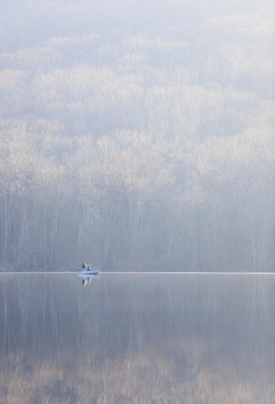 Small boat with a lone fisherman on a foggy, still pond at sunrise